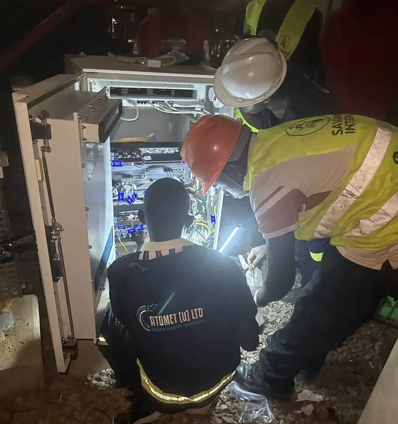Night work at a lit cabinet: an Atomet technician kneels at the equipment while a client engineer in a high-visibility vest holds a torch