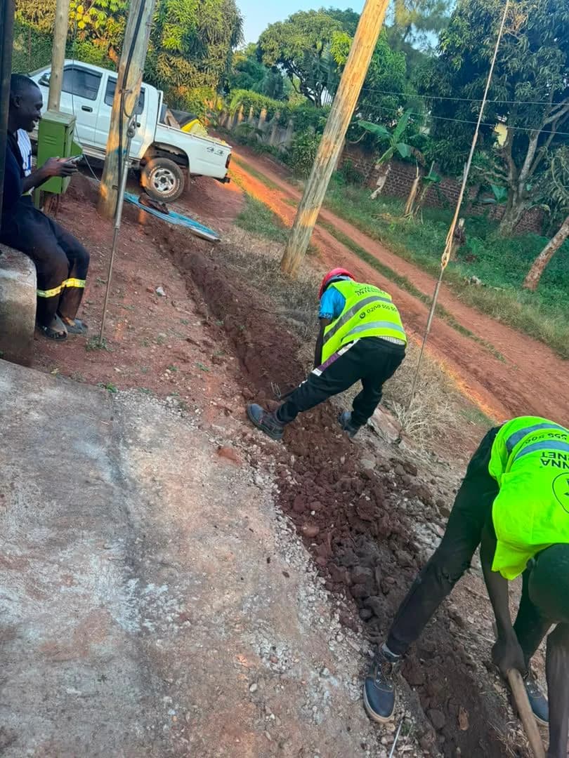Two workers in high-visibility vests digging a narrow trench along a red murram roadside for an underground fiber route