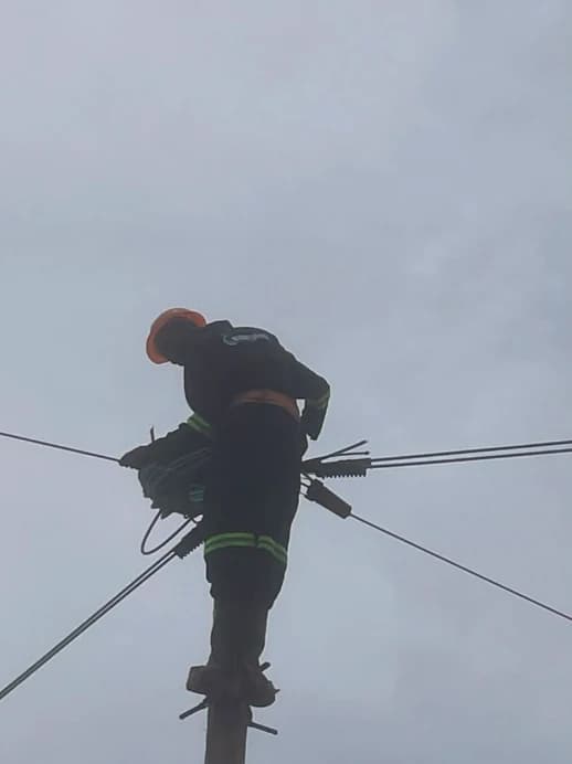 Atomet lineman in an orange hard hat and company coveralls standing at the top of a utility pole, fitting aerial fiber cables and clamps against an overcast sky
