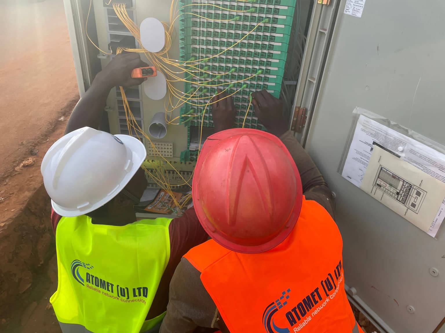 Two Atomet technicians in branded hi-vis vests, one in a white and one in a red hard hat, patching yellow fiber leads inside an open distribution cabinet