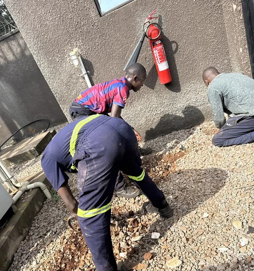 Crew in navy coveralls clearing gravel around a cabinet base to lay ducting beside a building wall