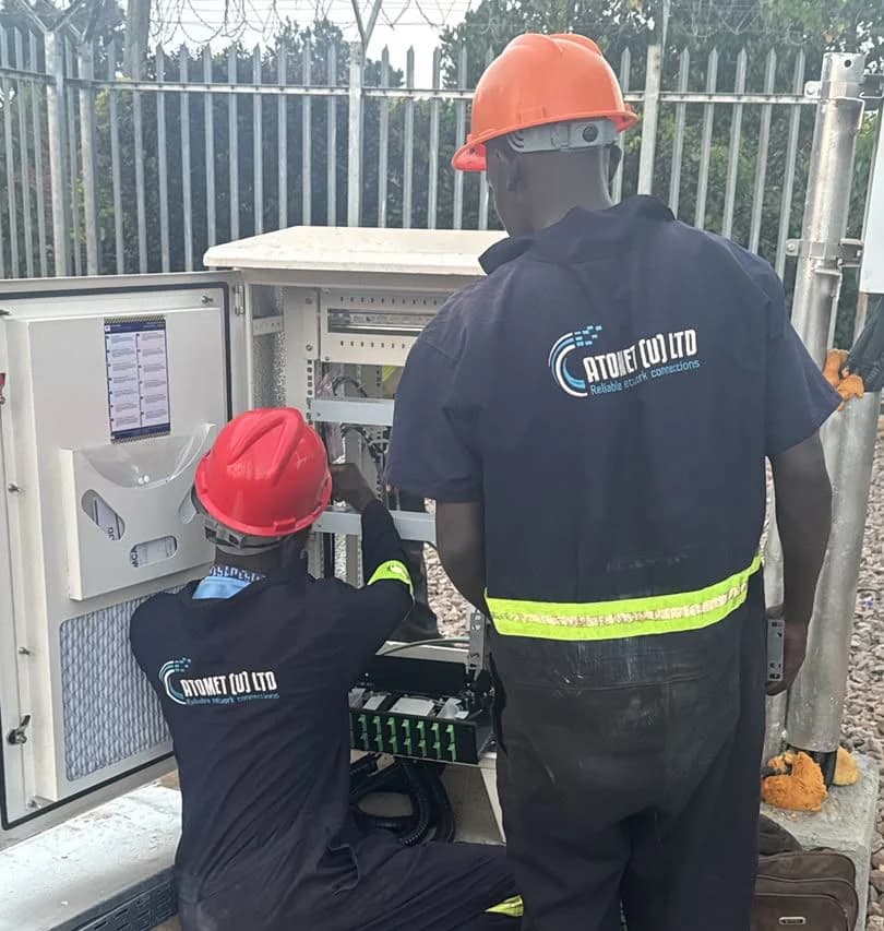 Two Atomet technicians in navy coveralls and hard hats commissioning equipment in an open cabinet, one kneeling at the splice tray