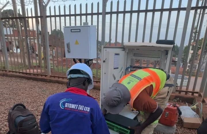 Atomet technician and a partner engineer kneeling at the base of an open cabinet inside a fenced compound