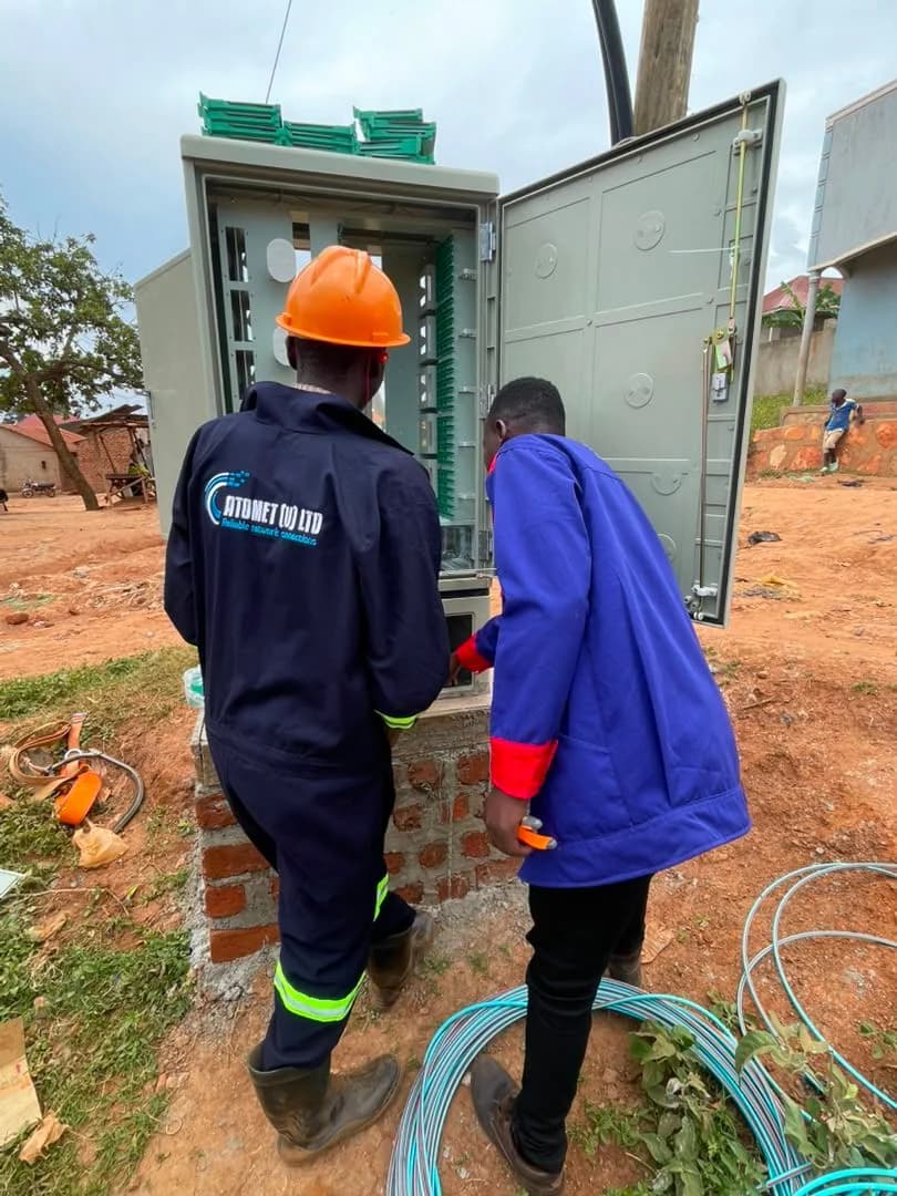 Two Atomet staff, one in an orange hard hat, at a tall green fiber distribution hub on a brick base with blue drop cable coiled on the ground