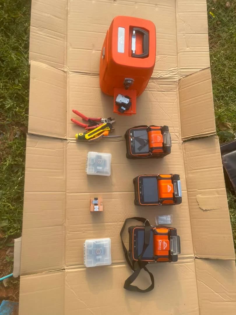 Orange fusion splicers, a cleaver, strippers and spare parts laid out on flattened cardboard on grass