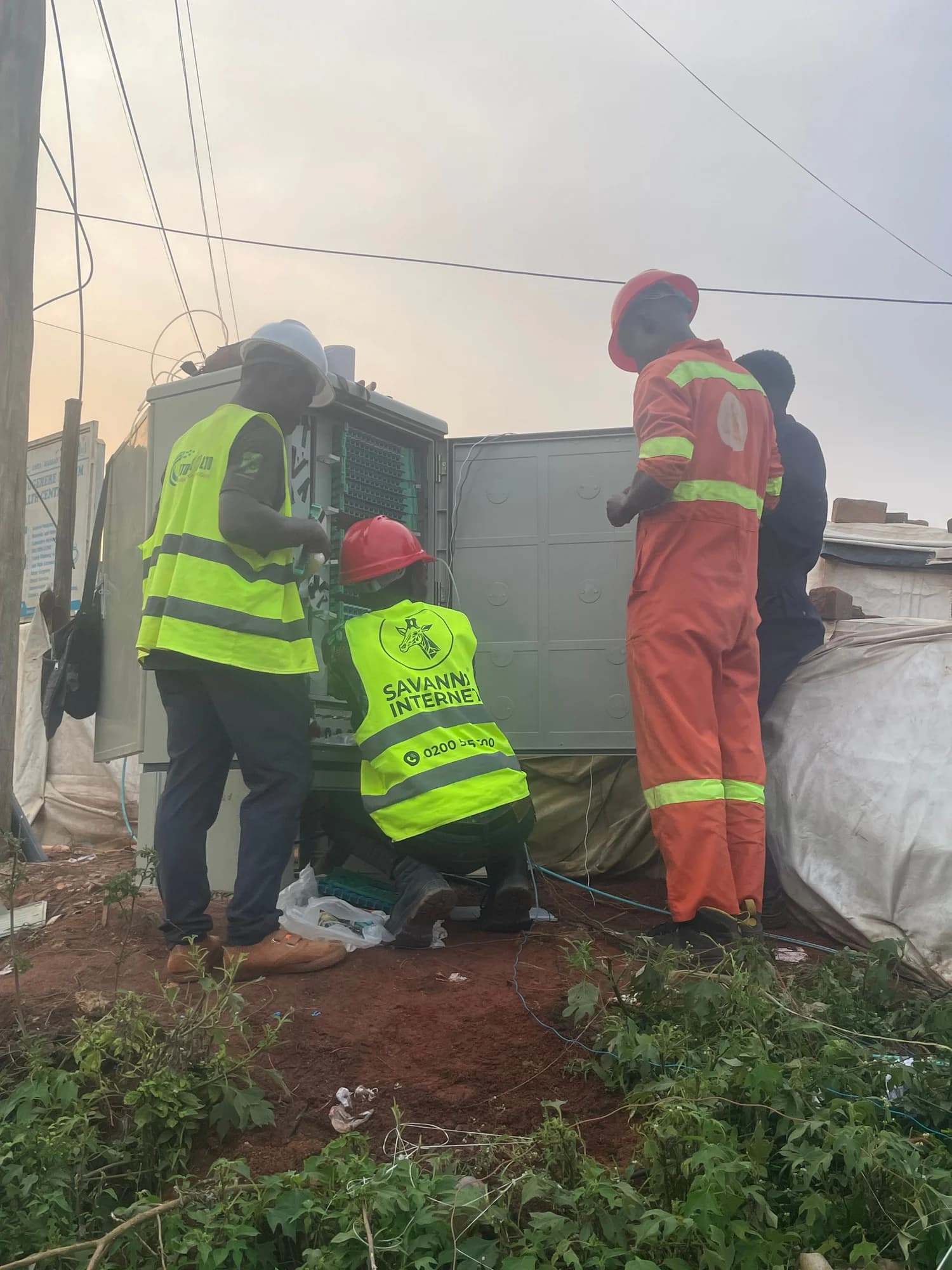Three people in hi-vis at an open roadside cabinet, one kneeling at the base, in early evening light