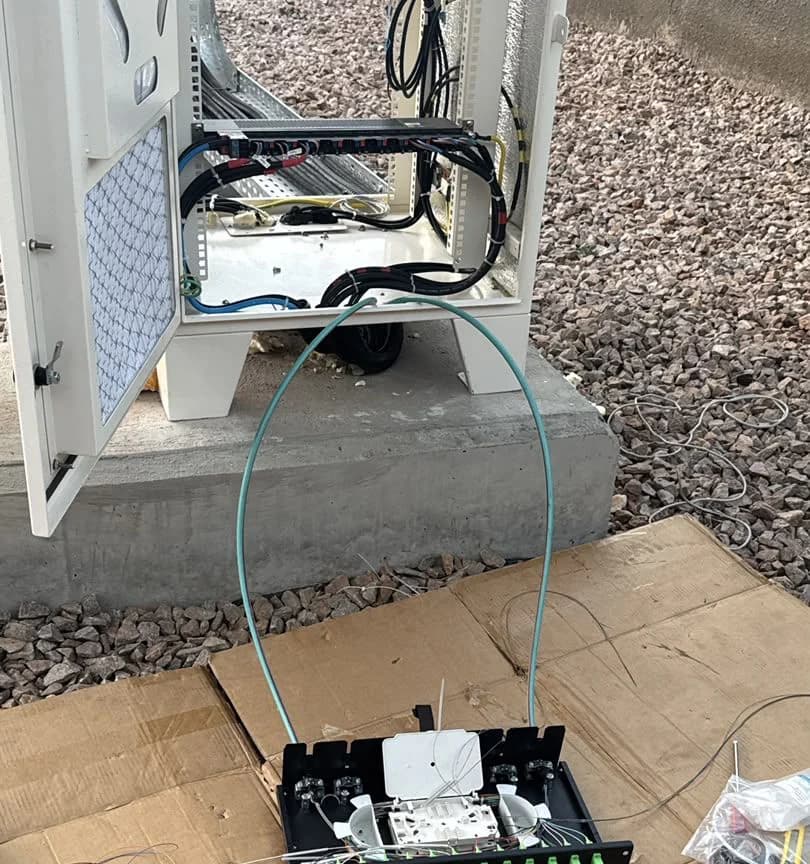 Open fiber termination box on cardboard at the foot of a cabinet, with a blue drop cable looping up into the cabinet