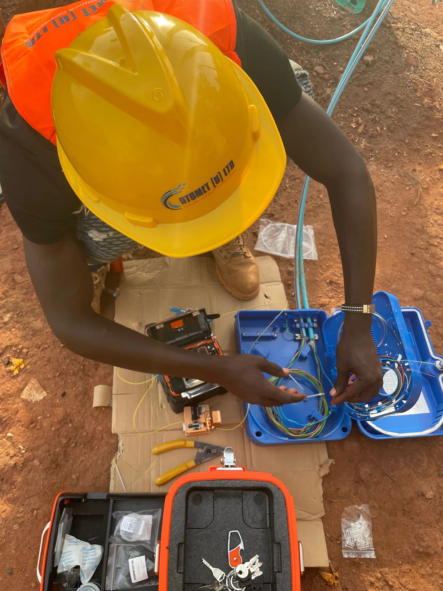 Technician in a yellow Atomet hard hat splicing fibers into a blue closure on a cardboard mat, splicer case open beside him