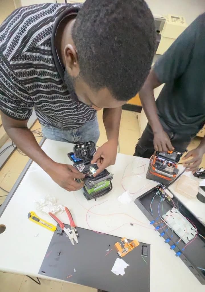 Two technicians at a white table preparing fiber in front of fusion splicers and a termination box