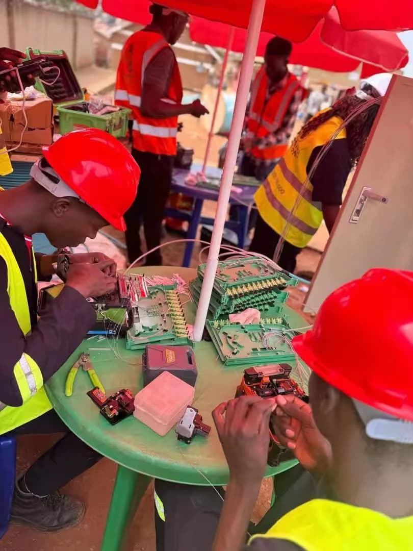 Technicians in red hard hats splicing fiber at a small table under a red umbrella beside a street cabinet