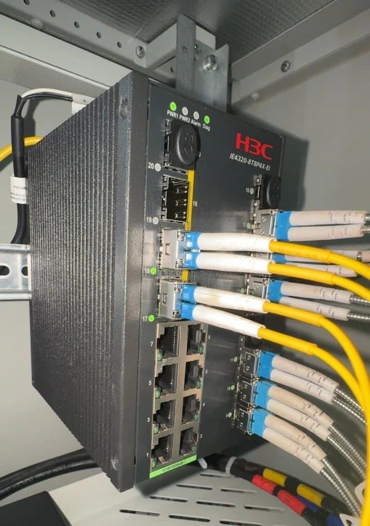 Industrial network switch mounted on a cabinet rail with yellow single-mode fiber patch leads plugged into its uplink ports