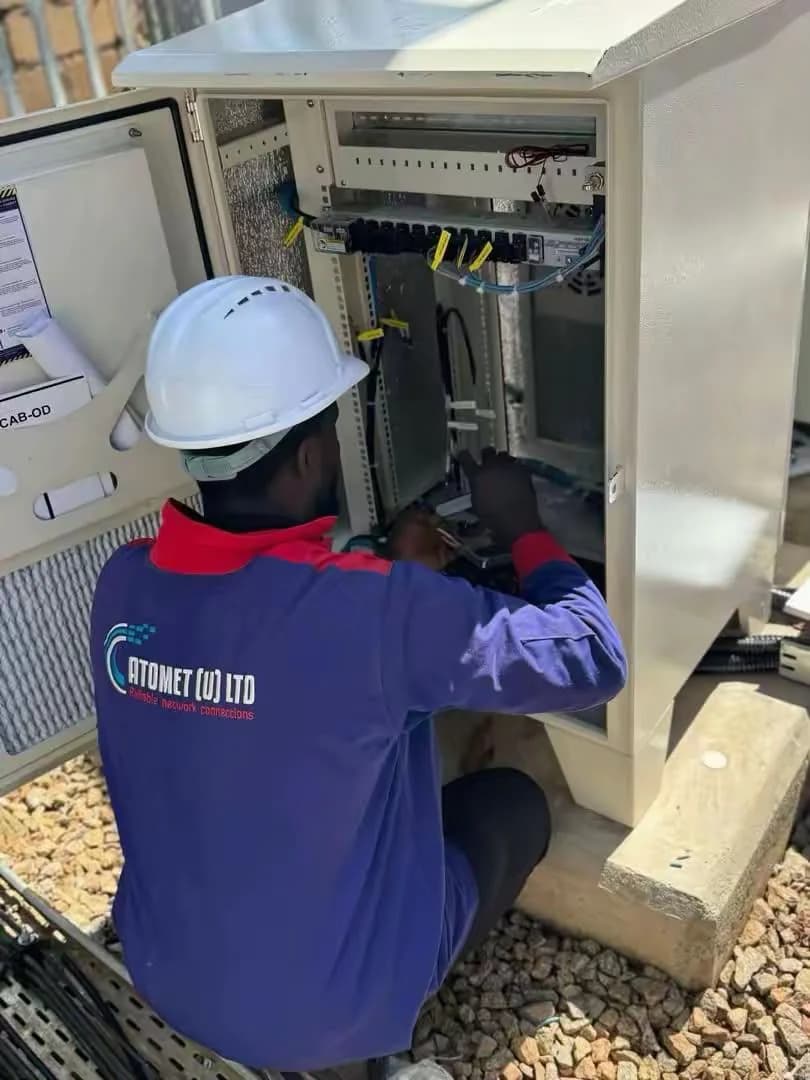 Atomet technician in a white hard hat and company jacket working inside an open roadside fiber cabinet, patch cords and splice trays visible